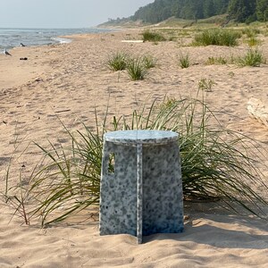 May include: A round, white stool with a speckled pattern sits on a sandy beach with green grass and a view of the ocean in the distance.