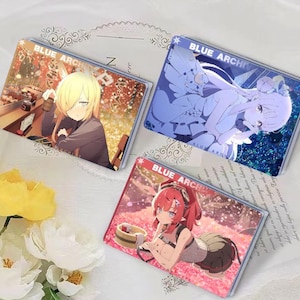 May include: Three Blue Archive character acrylic stands. The stands feature different characters from the game, including a blonde woman with a red bow, a woman with blue hair and a white dress, and a woman with red hair and a red and black outfit. The stands are all on a white background.