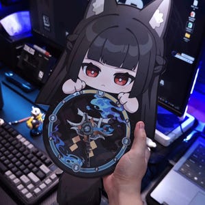 Puede incluir: Un personaje de dibujos animados con cabello negro y orejas de zorro sostiene un reloj redondo. La esfera del reloj presenta un diseño azul y dorado con los números 3, 6, 9 y 12. El personaje tiene ojos rojos y sostiene el reloj frente a una computadora.