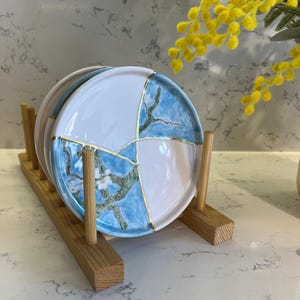 Può includere: Set di quattro sottobicchieri in ceramica blu e bianca con dettagli dorati. I sottobicchieri sono impilati su un supporto in legno.