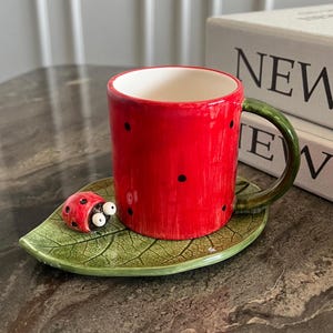 Peut inclure: Une tasse en céramique rouge avec des pois noirs, une anse verte et une soucoupe en forme de feuille. Une figurine de coccinelle est posée sur la soucoupe. Le mot "NEW" est partiellement visible en arrière-plan.