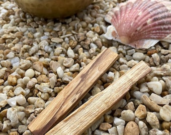 Puede incluir: Un cuenco tibetano dorado con una maza de madera, dos trozos rectangulares de madera de Palo Santo y una concha sobre un lecho de pequeños guijarros claros. El Palo Santo tiene un color marrón claro con vetas marrones oscuras.