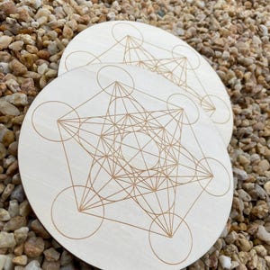 Peut inclure: Deux dessous de verre ronds en bois clair avec un motif géométrique du Cube de Métatron gravé en brun chaud. Les dessous de verre sont présentés sur un lit de petits galets clairs.