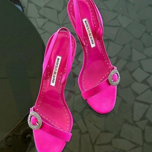 Puede incluir: Un par de tacones de satén rosa brillante con tiras traseras. Cada zapato presenta una hebilla plateada brillante y la marca "MANOLO BLAHNIK" impresa en una etiqueta blanca. Los tacones tienen un diseño delgado.