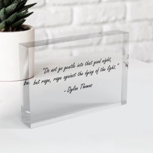 May include: A clear acrylic block with the quote "Do not go gentle into that good night, but rage, rage against the dying of the light." by Dylan Thomas. The block is rectangular and sits on a white surface.