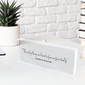May include: A clear acrylic block with the quote "The only thing we have to fear is fear itself" by Franklin D. Roosevelt. A small succulent in a white pot is to the left. A stack of books is on the right.