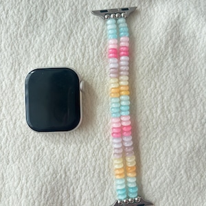 Puede incluir: Un Apple Watch plateado junto a una correa de reloj con cuentas. La correa está hecha de cuentas de colores pastel en tonos rosa, amarillo, azul y blanco. El reloj tiene una pantalla negra.