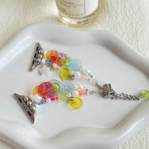 May include: A multi-coloured Apple Watch strap adorned with beads, buttons, and charms. The strap has silver-toned connectors and a chain clasp. The design incorporates clear, orange, yellow, green, and pink elements. The strap is on a white tray.