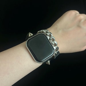 May include: A black smartwatch with a silver case and a black screen. The watch is paired with a silver chain-link band and black leather straps with silver cone studs. The watch is on a wrist.