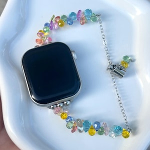 Op de afbeelding: Een smartwatch met een zwart rechthoekig scherm en een zilverkleurige metalen behuizing. De horlogeband bestaat uit veelkleurige, gefacetteerde, druppelvormige kralen in blauw, roze, geel en groen, verbonden door een zilverkleurige ketting.