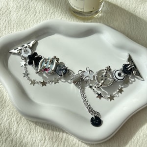 May include: A decorative watch band with silver-tone metal attachments and a chain. The band features black star-shaped charms, beads, and a dangling charm. The band is adorned with small silver stars and black and white circular accents.