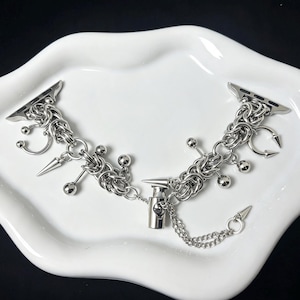 Könnte beinhalten: Ein silberfarbenes Uhrenarmband mit einem komplexen Kettengeflecht-Design. Das Armband ist mit mehreren hängenden Anhängern verziert, darunter kleine Kugeln, spitze Dornen und hufeisenförmige Elemente, die durch einen zylindrischen Verschluss verbunden sind.