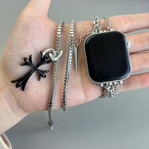 May include: A black cross pendant and a silver watch band with a black watch face are displayed. The watch band is connected to a silver chain. A silver spike charm hangs from the watch band. The items are displayed on a light gray surface.