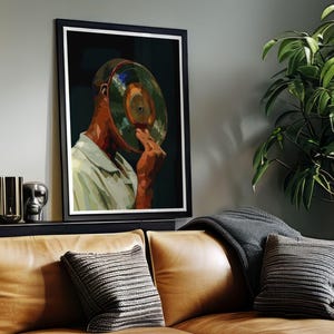 May include: Framed art print featuring a person with a vinyl record covering their face. The artwork has a dark background and a black frame. The person is wearing a light green shirt. The image is displayed above a brown leather couch.