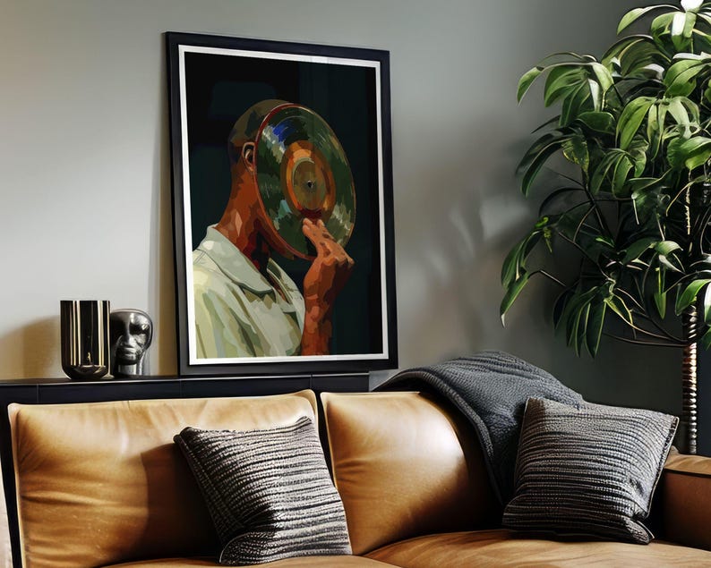 May include: Framed art print featuring a person with a vinyl record covering their face. The artwork has a dark background and a black frame. The person is wearing a light green shirt. The image is displayed above a brown leather couch.