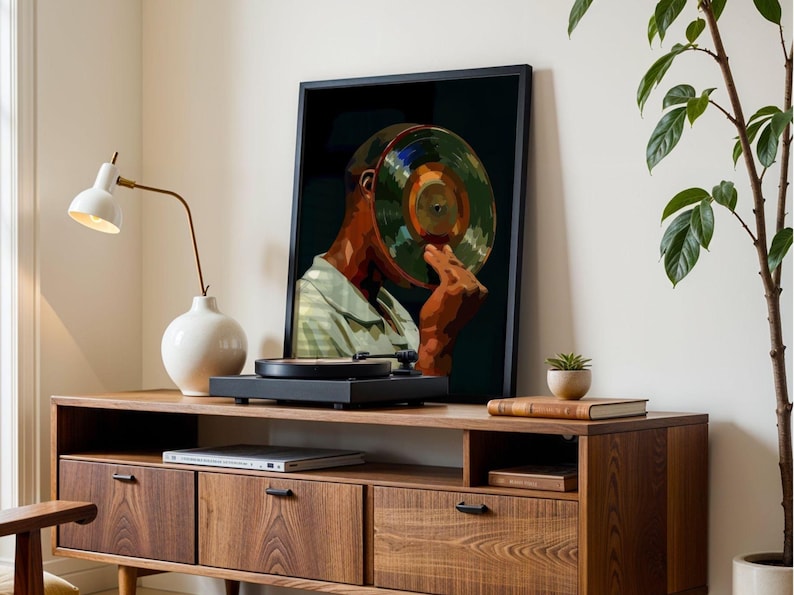 May include: A framed art print depicts a person with a record in place of a head. The artwork is displayed on a wooden cabinet with a record player. A white lamp and a white vase are also on the cabinet.