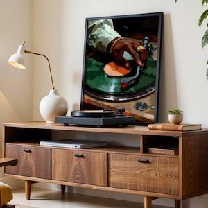Op de afbeelding: Een houten entertainmentcenter in vintage stijl met drie lades en een plank. Op de plank staat een zwarte draaitafel, en aan de muur erachter hangt een ingelijst afdruk van een hand die een plaat op een draaitafel aanpast. Een witte lamp met een gouden voet staat op de plank links van de draaitafel.