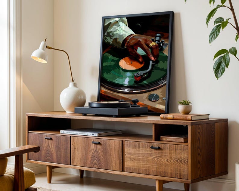Op de afbeelding: Een houten entertainmentcenter in vintage stijl met drie lades en een plank. Op de plank staat een zwarte draaitafel, en aan de muur erachter hangt een ingelijst afdruk van een hand die een plaat op een draaitafel aanpast. Een witte lamp met een gouden voet staat op de plank links van de draaitafel.