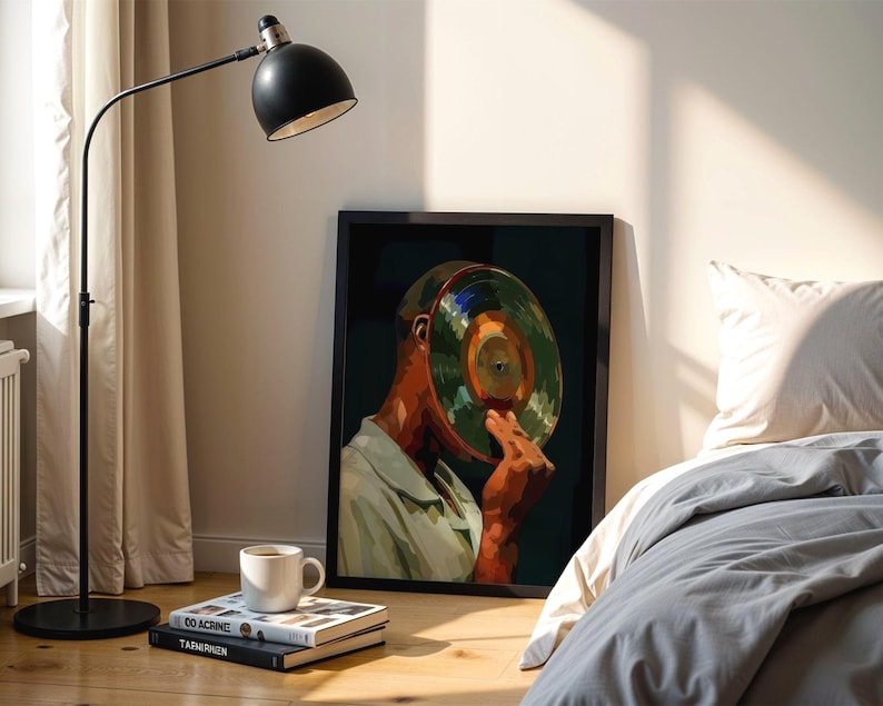 May include: A framed art print depicts a person holding a vinyl record to their face. The artwork features a color palette of green, orange, and brown. A black floor lamp and a white coffee cup on books are in the foreground.