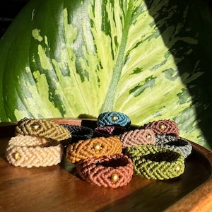 Könnte beinhalten: Eine Sammlung von Makramee-Ringen in verschiedenen Farben, darunter Beige, Olivgrün und Braun, jeweils mit einer kleinen goldenen Perle. Die Ringe werden auf einem Holztablett präsentiert, mit einem großen grün-weißen Blatt im Hintergrund.