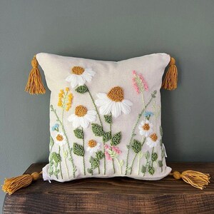 Peut inclure: Coussin carré crème brodé de marguerites blanches, de fleurs sauvages jaunes et roses et de feuilles vertes. Le coussin a des glands jaunes à chaque coin. Le coussin est posé sur une étagère en bois foncé.