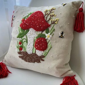 Peut inclure: Coussin beige avec broderie de champignons rouges, feuilles vertes et petites fleurs blanches. Le coussin est orné de pompons rouges et d'abeilles brodées. Le coussin est posé sur une chaise blanche.