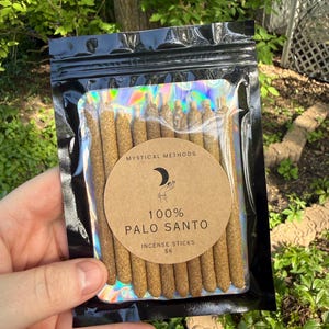 May include: A black, resealable bag containing multiple Palo Santo incense sticks. The bag has a holographic interior and a circular label that reads "100% Palo Santo Incense Sticks" with the brand name "Mystical Methods". The sticks are a light brown color.