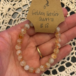 May include: A Golden Healer Quartz bracelet with round, translucent beads in shades of yellow and orange. The bracelet is strung on a thin, orange cord. A brown tag reads "Golden Healer Quartz".