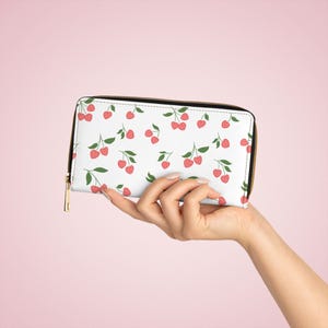Puede incluir: Cartera blanca con un estampado repetido de cerezas rosas y hojas verdes. La cartera tiene una cremallera negra y un tirador dorado. La cartera se sostiene en una mano sobre un fondo rosa.