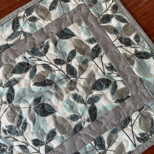 May include: A quilted table runner with a leaf pattern in shades of blue and gray. The runner has a gray border with a scalloped edge.