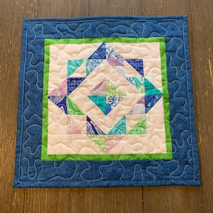 May include: A small quilted square with a blue and green geometric pattern. The center of the square is white and the pattern is made up of triangles. The square is surrounded by a blue border with a wavy design.