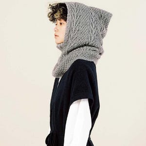 May include: A grey knitted hood and scarf set. The hood has a textured, open-weave design and frames the face. The scarf wraps around the neck. The person is wearing a dark blue vest over a white shirt.