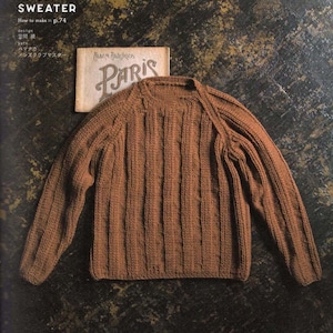 May include: A brown raglan sweater with vertical ribbed detailing. The image includes text that reads "RAGLAN SWEATER" and "PARIS". The sweater is displayed on a dark, textured surface, suggesting a vintage or handcrafted aesthetic.