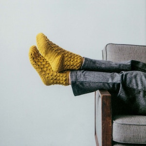 Può includere: Calzini all'uncinetto giallo senape con un motivo strutturato. I calzini sono indossati con pantaloni grigi e la persona è seduta in una poltrona grigia con braccioli in legno. I calzini sono il punto focale dell'immagine.