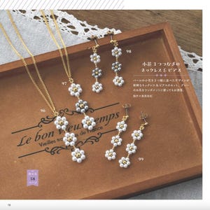 Op de afbeelding: Een ketting- en oorbellen set met bloemmotieven. De ketting heeft een gouden ketting met een hanger van witte en zilveren bloemen. De oorbellen hebben een vergelijkbaar ontwerp. De afbeelding staat op een houten dienblad.