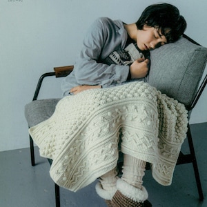 以下が含まれることがあります： クリーム色のテクスチャードかぎ針編みの毛布に覆われた人が椅子に座っています。毛布は詳細なパターンが特徴です。その人は靴下と茶色のかぎ針編みのスリッパを履いています。画像には上部に日本語のテキストがあります。