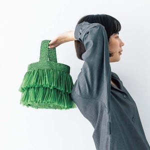 Puede incluir: Un bolso verde con flecos y un asa tejida. El bolso tiene un diseño en capas con múltiples capas de flecos. El bolso lo sostiene una persona que lleva una camisa gris de manga larga sobre un fondo blanco.
