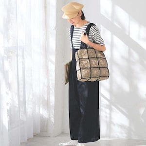 Puede incluir: Una mujer con gorra beige, camiseta a rayas y mono azul oscuro, lleva un bolso tote grande de crochet beige y negro. También lleva zapatillas blancas. La imagen está ambientada sobre un fondo blanco con una cortina transparente.