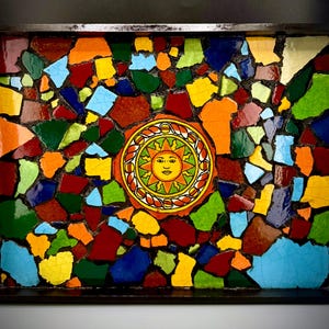 May include: A mosaic tile art piece featuring a central sun design surrounded by colorful, irregularly shaped tiles. The tiles are in shades of red, orange, yellow, green, blue, and brown, set within a dark frame. The sun has a smiling face.