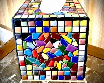May include: A decorative tissue box with a mosaic design. The box is square with a black border and a colorful mosaic pattern of various shapes and colors. A white tissue is pulled from the top.