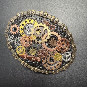 May include: An oval-shaped steampunk-style belt buckle featuring an intricate arrangement of gears in various sizes and metallic colors, including gold, copper, and silver. The gears are set against a textured black background and framed by a chain-like border.