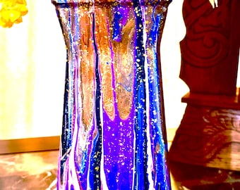 May include: A tall, hand-painted vase with a unique abstract design. The vase features a blend of blue, purple, gold, and white paint, creating a dripped effect. The vase is filled with colorful flowers, adding a vibrant touch to the composition.