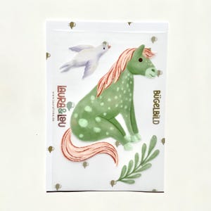 Könnte beinhalten: Grün-rosa Pferde-Bügeleisen mit weißen Punkten. Das Patch ist von kleinen braunen Schafen und einem grünen Blatt umgeben. Der Text "Bügelbild" ist in Gold auf der rechten Seite des Patches gedruckt.