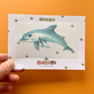 Könnte beinhalten: Ein blau-weißer Delfin-Aufbügler. Der Aufnäher ist rechteckig mit weißem Rand und kleinen braunen Vogelillustrationen. Der Text "BÜGELBILD" steht oben und "LAURA & LOU" unten.