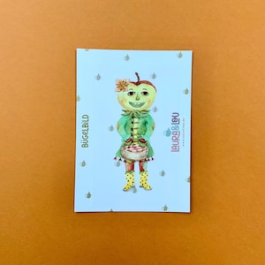 Könnte beinhalten: Eine skurrile Illustration einer apfelköpfigen Figur in einem grünen Mantel und gelben Stiefeln, die einen Korb trägt. Der Hintergrund ist hellblau mit kleinen Teekannenmustern. Die Texte "BÜGELBILD" und "LAURA & LOU" sind sichtbar.