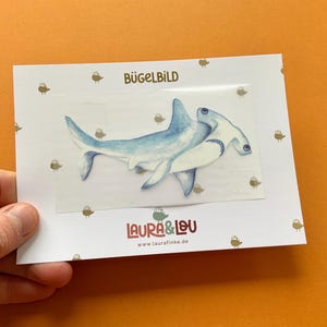 Könnte beinhalten: Bügelbild mit einer Illustration eines blauen und weißen Hammerhais. Das Bügelbild ist mit "Bügelbild" beschriftet und enthält die Website "www.laurafinke.de".