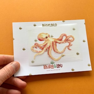 Könnte beinhalten: Ein Aquarell-Oktopus-Design auf einer weißen Karte mit dem Text "BÜGELBILD" und "LAURA & LOU". Der Oktopus ist in Orange- und Rosatönen gehalten. Die Karte hat eine transparente Folie und kleine Vogelillustrationen.