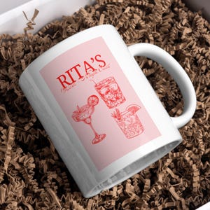 Puede incluir: Taza de cerámica blanca con fondo rosa y texto rojo que dice "Rita's Cocktails - Original Cocktails". La taza presenta tres dibujos de línea roja de cócteles.