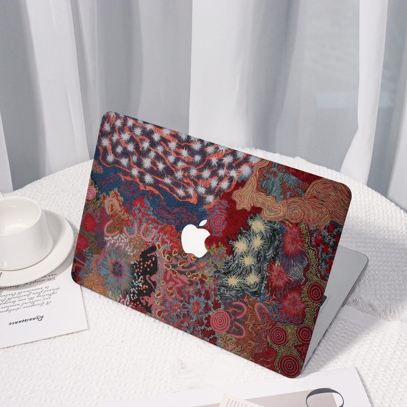 May include: A laptop computer with a colorful, patterned case. The case features a complex design with red, blue, and yellow hues, and a white Apple logo. The laptop is open, and a white coffee cup and saucer are visible.