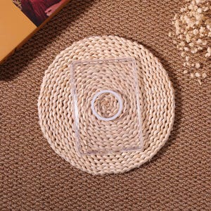 May include: A clear, rectangular phone case with a circular, white ring in the center. The case is placed on a round, woven straw mat, set against a brown, textured background. The overall aesthetic is natural and minimalist.